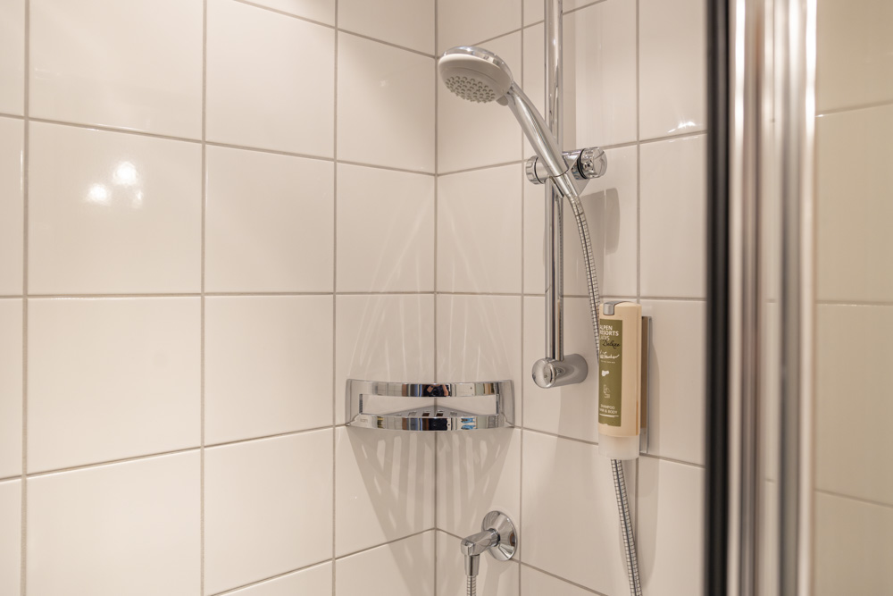 Eine moderne Dusche im Apartment für 4–6 Personen Serfaus-Fiss-Ladis verfügt über weiß geflieste Wände und einen silbernen verstellbaren Duschkopf an einer vertikalen Stange, komplett mit Seifenspender und kleinem Metallregal für Toilettenartikel, und bietet Komfort und Stil.