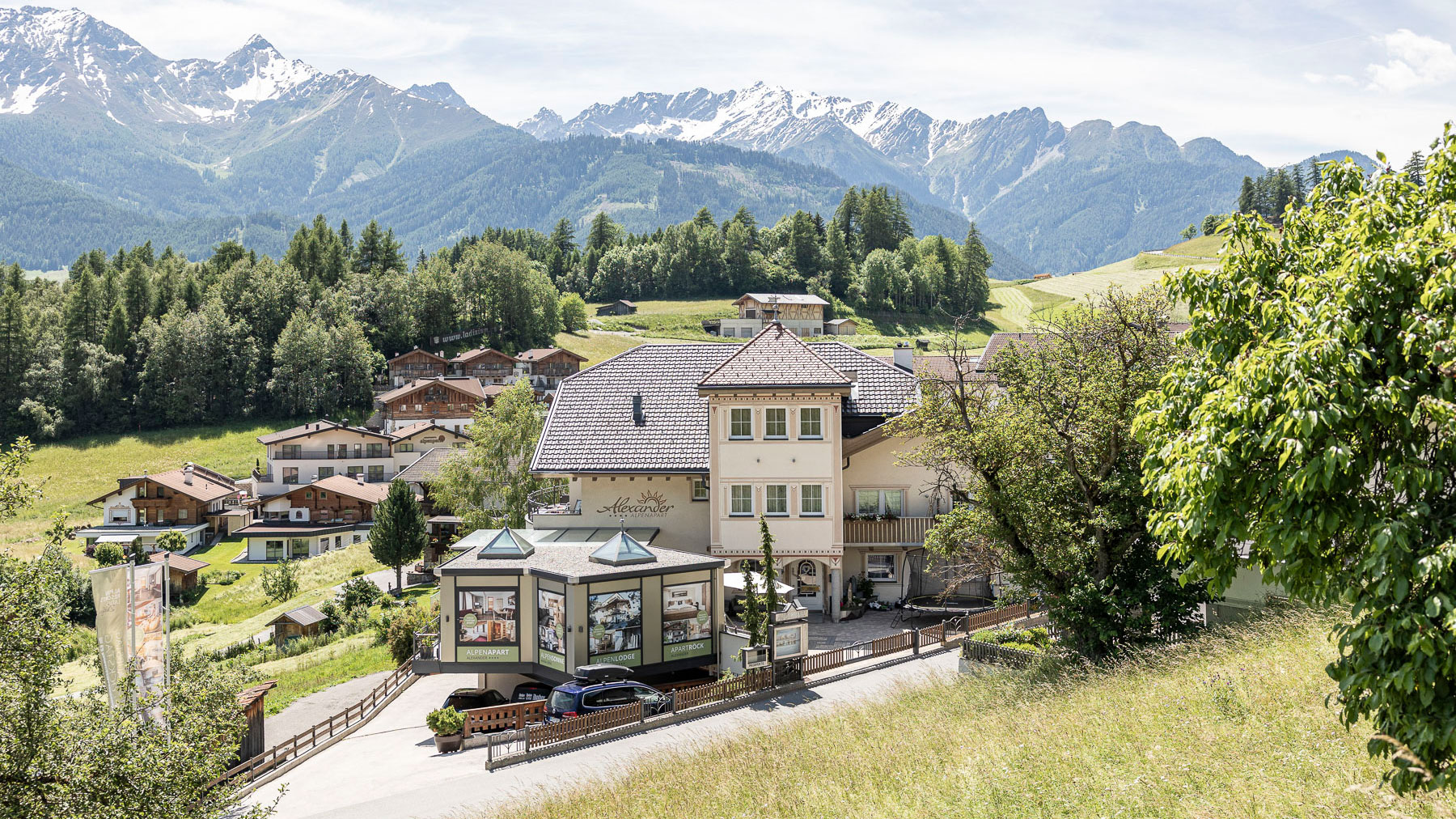 Eingebettet in ein üppiges grünes Tal bietet dieses malerische Dorf des Familienurlaubs Serfaus-Fiss-Ladis traditionelle alpine Gebäude. Im Hintergrund ragen schneebedeckte Berge unter einem klaren blauen Himmel auf, während Bäume und Grashänge die Gegend umhüllen und eine friedliche, malerische Umgebung schaffen.