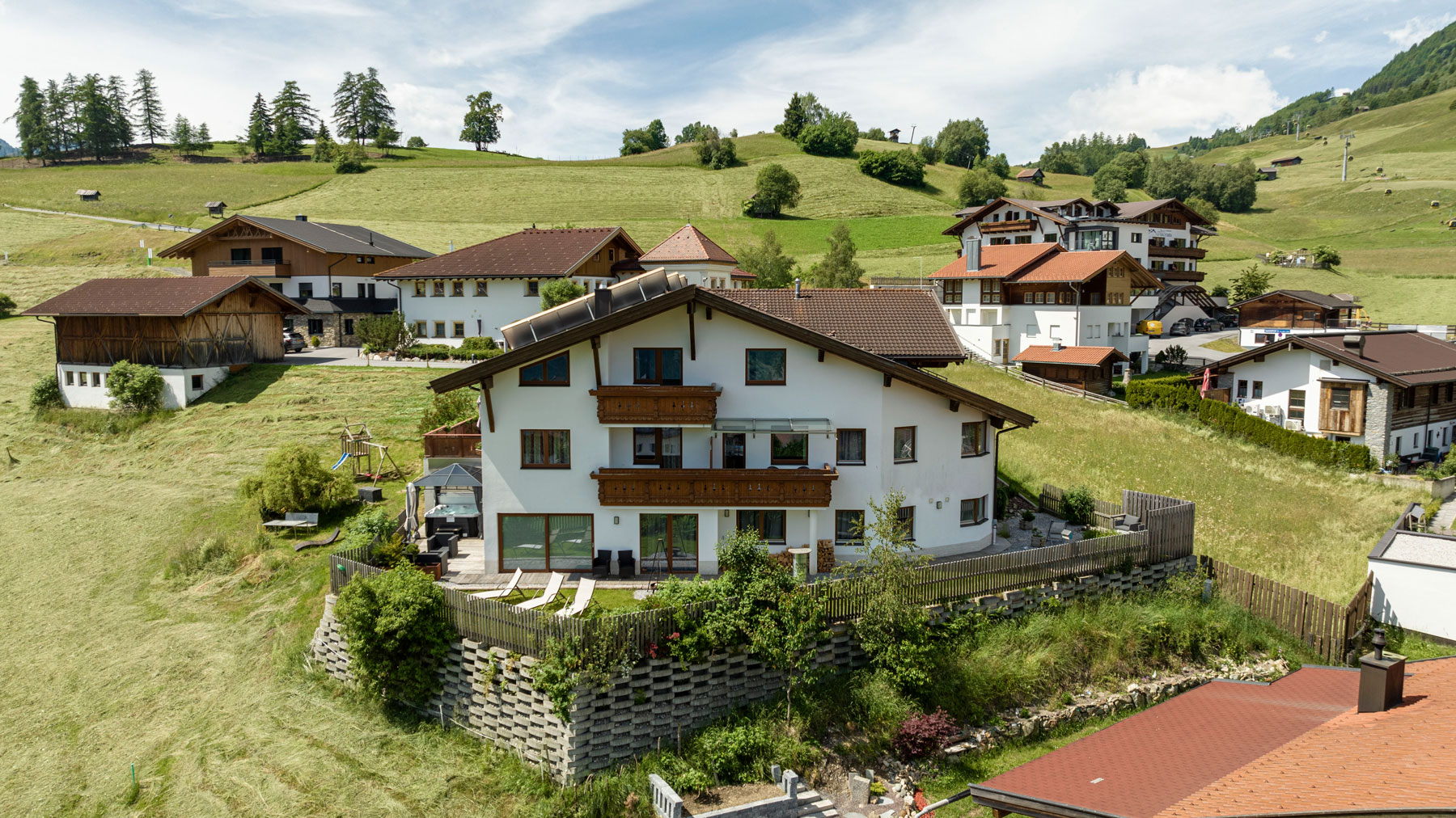 Eine Ansammlung moderner alpiner Chalets und Apartments liegt an einem grünen Berghang in der Nähe der Bergbahn in Ladis. Diese charmanten Gebäude verfügen über weiße Fassaden mit Holzakzenten und schrägen Dächern, umgeben von Grasfeldern, Bäumen und sanften Hügeln unter einem teilweise bewölkten Himmel.