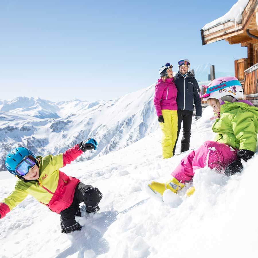 Zwei Kinder spielen im Schnee, während zwei Erwachsene, alle in Skikleidung, auf einem verschneiten Berg zusehen.