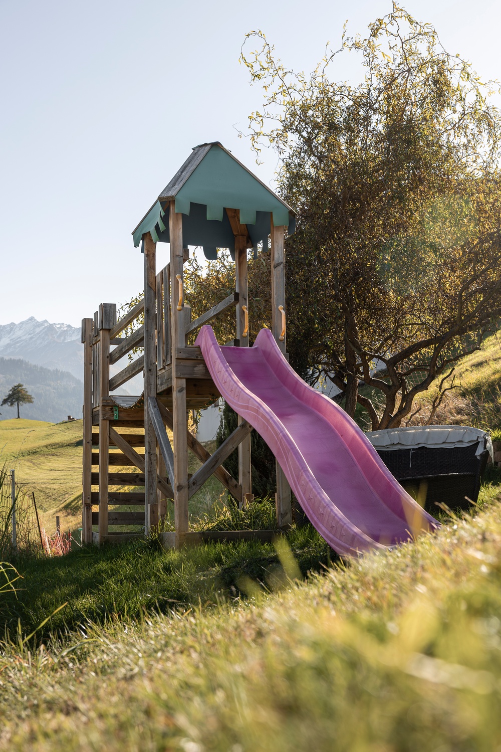 Holzspielgerät mit rosa Rutsche in einem grasbewachsenen Außenbereich mit Bäumen und Bergen.