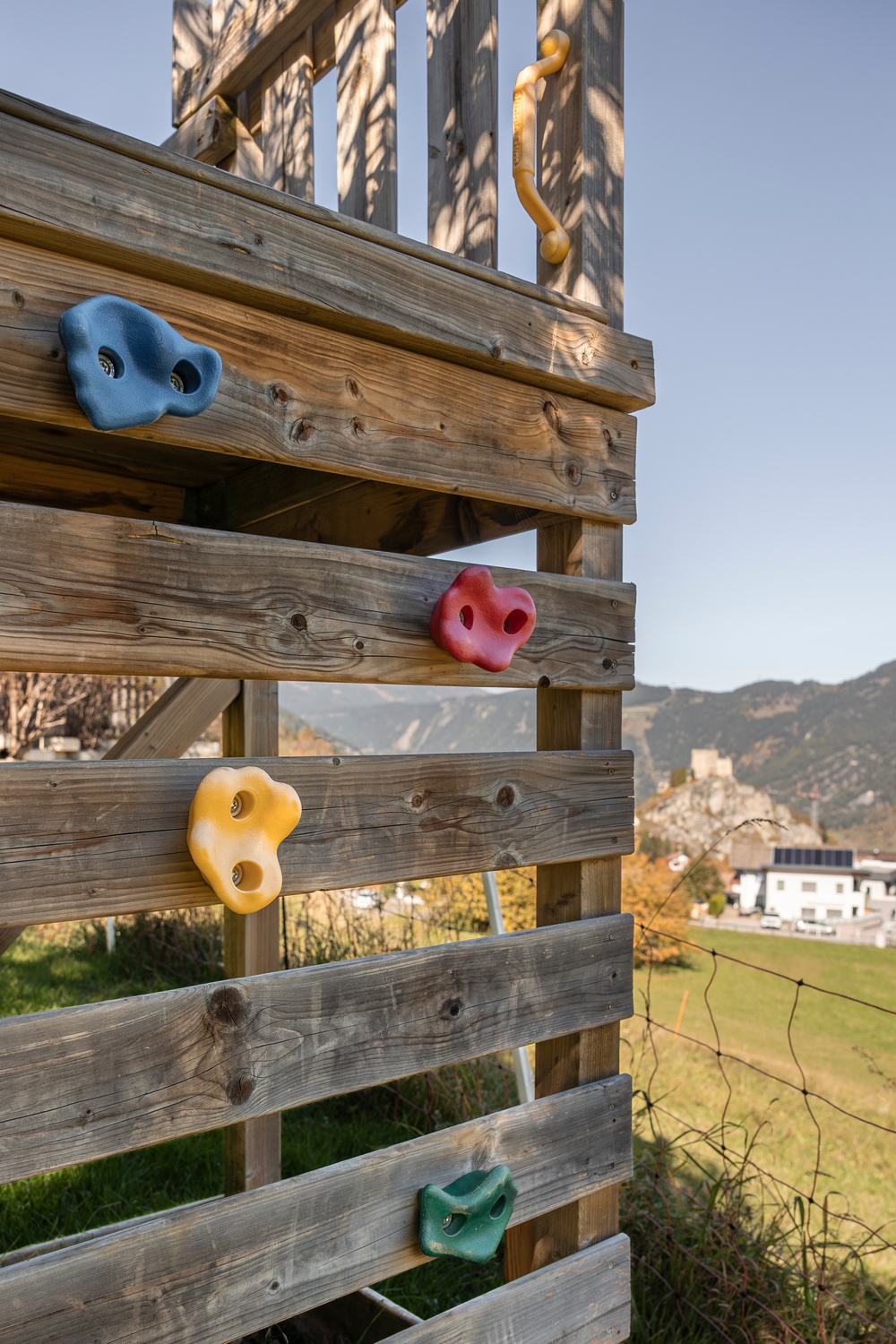 Hölzerne Kletterwand mit bunten Griffen, eine Wiese und Berge im Hintergrund.