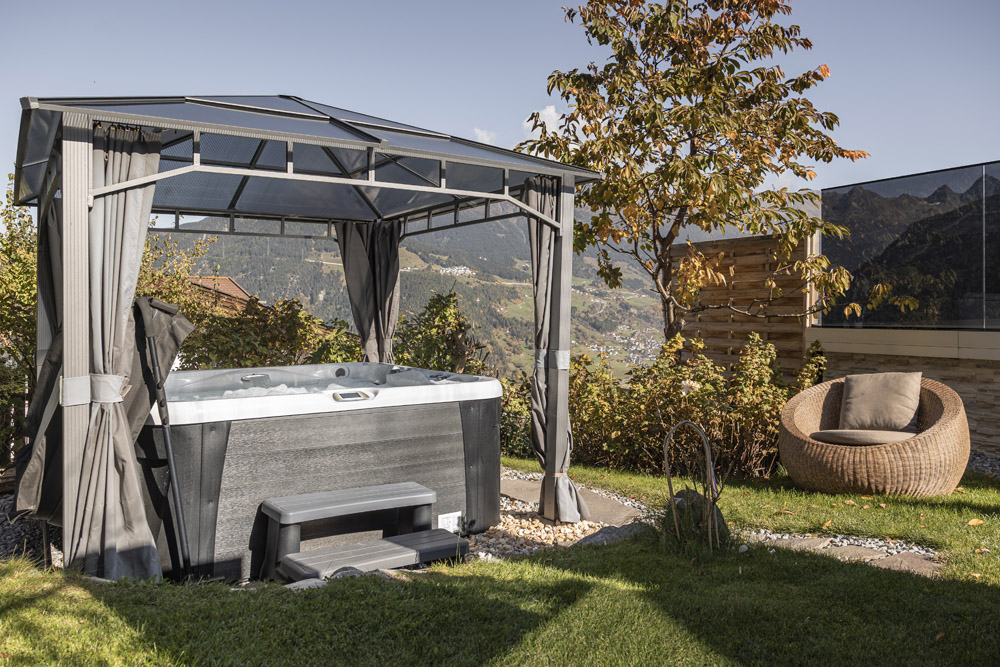 Ein Whirlpool unter einem Pavillon in einem Garten mit Korbstuhl und Bergblick.