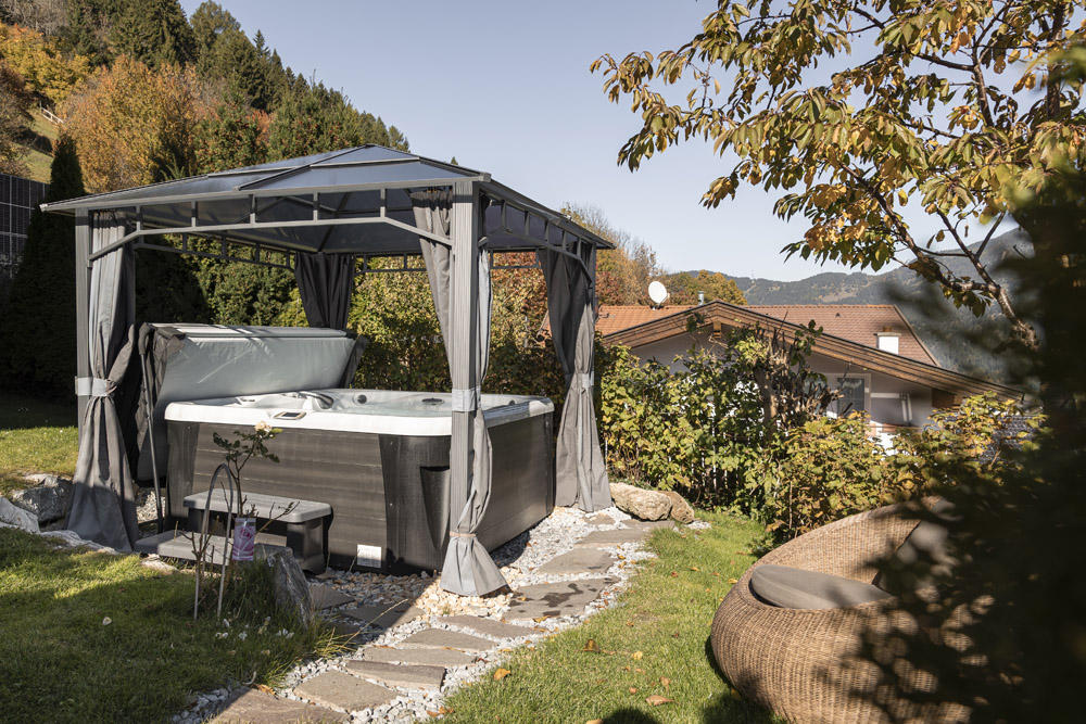 Whirlpool im Freien unter einem Pavillon in einem Garten, umgeben von Grün und Herbstbäumen.