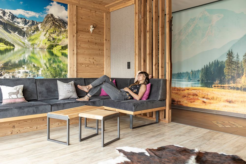 Eine Frau, die auf einem grauen Sofa in einem gemütlichen Zimmer mit Holzwänden und malerischen Bergwandbildern entspannt.