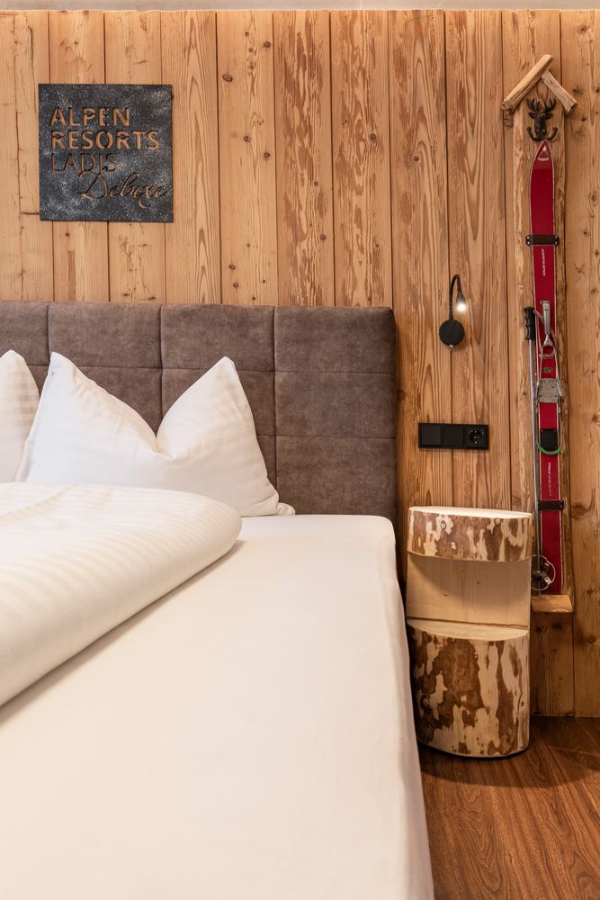 Ein gemütliches Bett mit weißer Bettwäsche, rustikale Holzwände, Skier an Haken und ein Nachttisch aus einem Baumstumpf.
