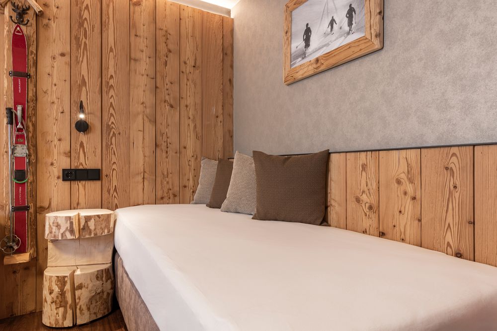 Gemütliches Holzzimmer mit einem Bett, Kissen, Skiern, einem Tisch aus Baumstamm und einem gerahmten Skifoto an der Wand.