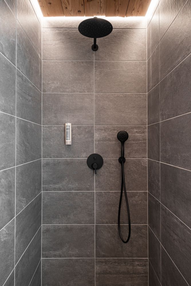 Moderne, mit grauen Fliesen ausgekleidete Dusche mit schwarzen Armaturen und einer Holzdecke mit Einbauleuchten.