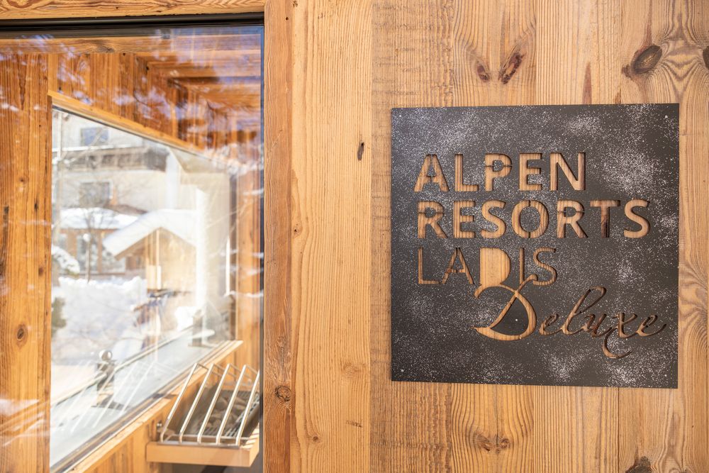 Ein Schild mit der Aufschrift „ALPEN RESORTS LADIS Deluxe“ an einer Holzwand neben einem Fenster, draußen liegt Schnee.