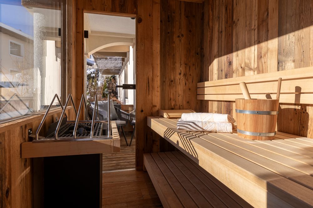 Innenansicht einer Holzsauna mit einem Eimer, einer Schöpfkelle, Handtüchern und Sonnenlicht, das durch ein Fenster hereinströmt.